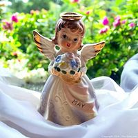 Front view of the angel figurine holding a basket of pastel flowers with "MAY" sash visible, set against a garden background.