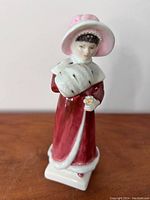 Front view of Royal Doulton Sophie figurine in red coat, white fur, and pink hat on wooden surface