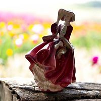 Front view of Royal Doulton porcelain figurine depicting a woman in a red dress and wide-brimmed hat, posed with one hand holding the hat and the other lifting part of the dress.