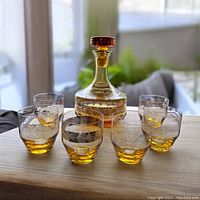 Full view of the amber glass decanter with stopper in the center surrounded by six matching drinking glasses on a wooden surface, all featuring embossed decorative patterns.