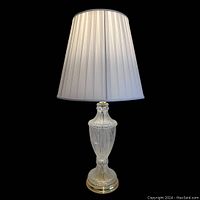 Full view of the lamp showing the crystal body, brass base, and white pleated lampshade.