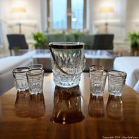 Photo showing six vintage crystal glasses with detailed cut glass patterns arranged around a matching crystal ice bucket with a silver handle and rim on a wooden table, focused in a living room setting.