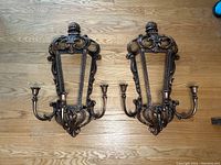 Pair of vintage brass-like wall sconces with ornate scrollwork and two candle holders each, side by side on wooden floor.