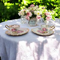 Two tea cup and sandwich plate sets placed on white tablecloth outdoors with floral centerpiece in background. Pink set on left, blue set on right.