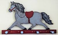 Full view of the hand painted wooden horse-shaped rack mounted on a reddish-brown base with 4 round pegs painted light blue. Horse painted in grey with black mane and tail, red saddle and bridle, signed 'Cole' on leg.