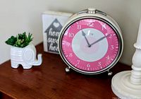 Large silver metal framed round alarm clock with bright pink and white concentric circles dial, white numerals, standing on metal feet placed on a wood surface alongside white elephant planter and decor sign.