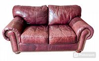 Front view of burgundy leather loveseat showing brass tack details along rolled arm fronts and removable cushions with visible patina and wear.