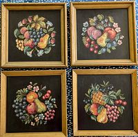 Set of four framed needlepoint fruit and flower artworks arranged on a blue floral background. Each artwork shows a unique arrangement of fruits and flowers on black fabric within gold painted frames.