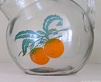 Close-up photo of the side of the glass pitcher showing the painted orange fruit and leaves design on clear glass.