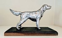Side profile of metallic long-haired pointer dog figurine on black rectangular wooden base.