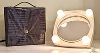 Photo of black vinyl carrying case with handle and clasp, next to vanity mirror with bulbs around mirror edge, for portability and storage indication.