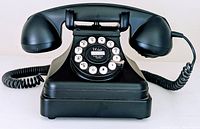 Photo showing front and side view of vintage black rotary dial telephone with handset on cradle and coiled cord