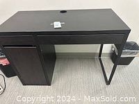 Full frontal view of black wood grain desk showing storage drawer and cabinet on left side, metal legs, and cable management hole on top.