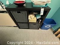 Front wide view of black wooden storage shelf with three black woven wicker baskets inserted in the lower compartments, a red bowl on top, and papers stacked on top.