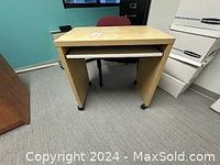Front view of the light wood mini desk showing the top surface, side panels, and keyboard shelf, positioned in an office setting with carpet and surrounding furniture.