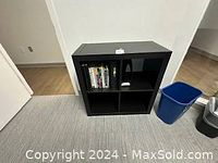 Front view of dark wood grain storage shelves with four compartments, some containing books and objects, next to a blue trash bin.