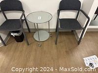 Full view of the glass top rolling table flanked by two gray chairs on a wood floor.