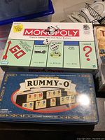 Monopoly and Rummy-O games stacked together showing box covers: Monopoly with green background and classic property names, Rummy-O metal tin with tile numbers.