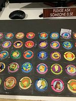 View of multiple Simpsons character pogs arranged in rows under plastic storage sheets on a table showing colorful cartoon images and names.
