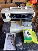 Front view of the Singer sewing machine with foot pedal and accessory container placed nearby on a black surface, along with the instruction manual.