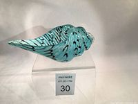 Side angled view of turquoise and black hand blown glass conch shell vase on a clear stand, showing intricate pattern and smooth glossy finish