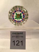 Front view of faceted glass paperweight showing green leaves and colorful millefiori flowers encircled by blue and pink canes