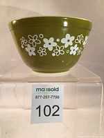 Front view of olive green Pyrex mixing bowl with white daisy flower designs on the exterior, displayed on clear stand with lot number 102