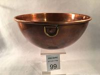 Side view of the copper bowl showing its domed shape and brass hanging loop, placed on a clear stand with lot number tag.