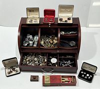 Full view of wooden box with compartments and drawer holding assorted men's accessories including watches, cufflinks, rings, and tie clips. Also shows some items in presentation boxes alongside the box.