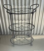 Full view of the vintage Bowery 3 tier oval glass beverage cart on wheels with clear glass shelves and black metal frame