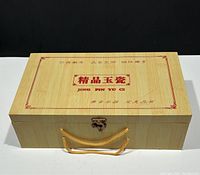 Closed natural wood box with printed red Chinese characters and English text 'JING PIN YU CI'. Gold cord handle and metal latch closure.