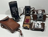 Collection of four vintage cameras including Ansco Karomat folding camera, Voigtlander folding camera with carrying cases, Riken Ricoh 35, and Tokyo Primo Jr. twin lens reflex camera with cases arranged for display.