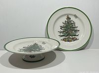 Two Copeland Spode Christmas Tree plates shown together, one flat and one footed cake plate, both with green rims and colorful Christmas tree designs.