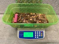 Green plastic container filled with mixed pennies and a pink penny wrapper on a digital scale showing weight 7.034 pounds.
