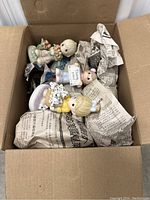 Box top open showing three child figurines with protective packing paper inside a cardboard box.