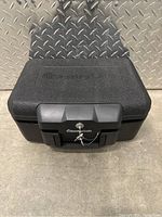 Closed view of black SentrySafe portable lockbox safe with key lock on front latch, placed on floor with diamond plate metal wall background.