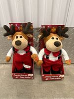 Front view of two dancing reindeer plush toys in original festive packaging, showing clothing and facial features.