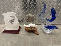 Four dolphin sculptures arranged side by side on a surface with diamond plate background showing varied materials and styles.