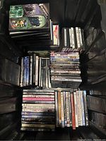 Top-down view of black storage bin filled with assorted DVDs in cases stacked vertically and horizontally showing some titles.
