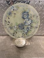 Front view of a round glass plate with floral hand-painted design in blue and white on a frosted glass background, positioned on a holder against a metal textured backdrop.