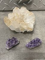 Three gemstone mineral specimens placed on a metal surface, showing one large clear quartz cluster and two smaller amethyst clusters.