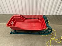 Red plastic kids sled with green metal frame and yellow pull rope sitting on diamond plate metal surface indoors.