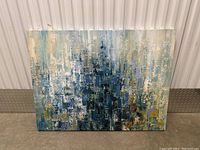 Large rectangular abstract canvas painting resting on floor against a corrugated white background, showing textured layered paint in blue, white, gold, and green hues.