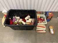 Top-down view of a lot of assorted candles and related items inside a large black storage bin, with some candles and boxes surrounding the bin.