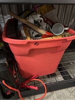Plastic red bin filled with assorted tools and boxed, bagged miscellaneous items including a wooden stick with red funnel, a tool with red handles partially outside bin