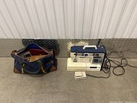 Sewing machine alongside a navy blue carrying bag containing sewing supplies, displayed on a gray floor.