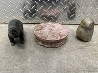 Three stone sculptures: a small black bear, a circular scalloped reddish stone base, and a small owl with engraved eyes. Background is a concrete floor and metal diamond plate.