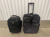 Photo showing the two suitcases side by side on a concrete floor with metal wall base. Gray suitcase on right and black HEYS suitcase on left.