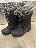 Pair of black women's winter boots with faux fur collar, lace-up front, and quilted fabric upper, shown side angle on metal textured surface.