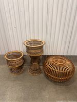 Full lot shot showing two rattan pedestal baskets and one round rattan ottoman on concrete floor.